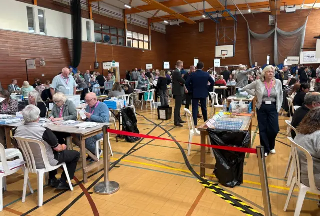 People in a sports hall sitting at table counting ballots