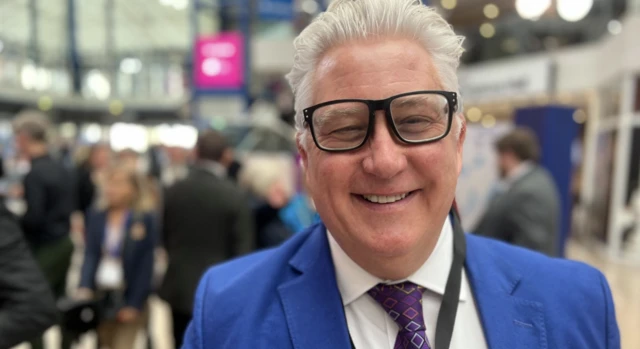 Simon Bentley with silver hair, black rimmed glasses and a bight blue blazer smiling at the camera