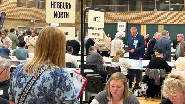 The election count in a brown and green sports hall. There are multiple rows of tables, lined with people ruffling through ballot papers. with white signs erected above them. In the foreground, there are half a dozen people looking on at those counting.