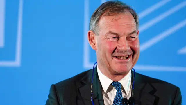 Rupert Lowe MP smiling, with a blue backdrop behind him.