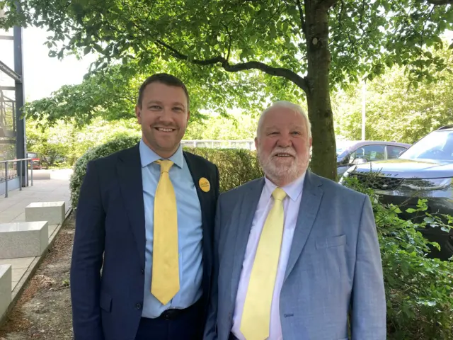 Henry on the left and John on the right smile at the camera. Both wear yellow ties.