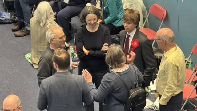 Six people are standing in a circle and are talking between each other. On the right is a row of red chairs where a few people are sitting on them.
