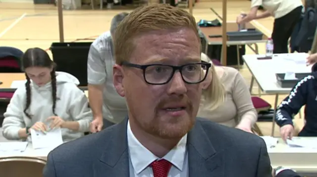 Jonathan Brash is standing in front of people who are rifling through ballot papers. He has short ginger hair and black glasses. He is wearing a blue suit with a red tie. He has a concerned look on his face as he speaks with reporters.