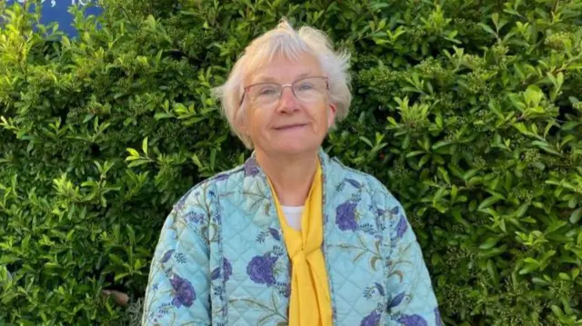 Inga Lockington is standing in front of the BBC Radio Suffolk building you can see a hedge to the right in the background and to the left there is a BBC Radio Suffolk sign. Inga is wearing patterned purple and teal padded coat with a yellow scarf. She is smiling at the camera.