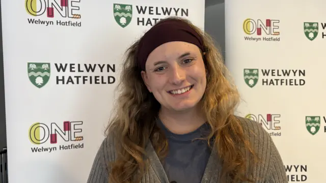 Sandrini Bonfante with long brown hair wearing a blue top and grey sweater smiling at the camera in front of Welwyn Hatfield council display boards.