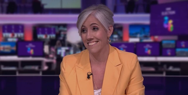 Daisy Cooper wearing a yellow blazer while sat in the BBC news studios. She is smiling looking to the left of the camera