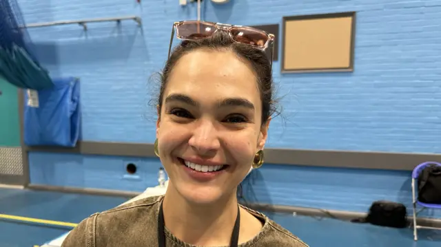 A woman smiles at the camera. She has dark hair that has been tied back and wears a light denim top and gold earrings.
