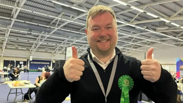 Martin Osborne is smiling brightly with both thumbs raised. He is wearing a green rosette and dark three-quarter zip fleece. People are sitting on tables dotted around the sports hall behind him.
