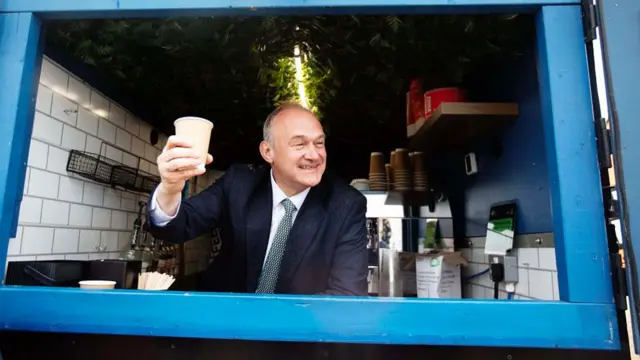 Ed Davey holds up a cup of coffee in celebration - he is smiling
