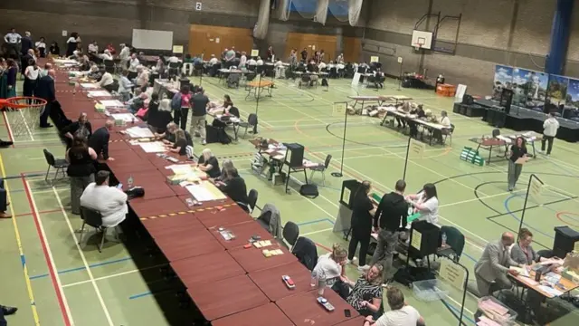 A large brown table is running across a building on the left. People are sat either side on chairs. Elsewhere in the room, there are tables spread across different areas and a stage.