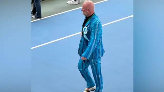 A man walking through the sports hall wearing a striking blue, striped suit and a turquoise rosette for Reform UK. He has blading white hair and a beard and is wearing white shoes.