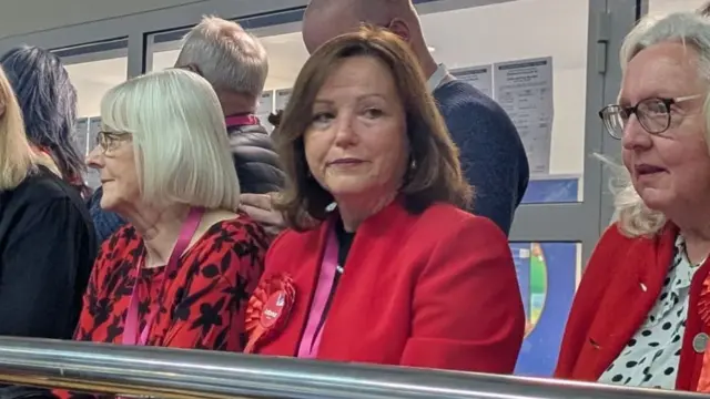 Karen Clark is sitting and looking off the the right with a dejected look on her face. She has mid-length brown hair and is wearing a red jacket and red rosette. There are people sat on either side of her.