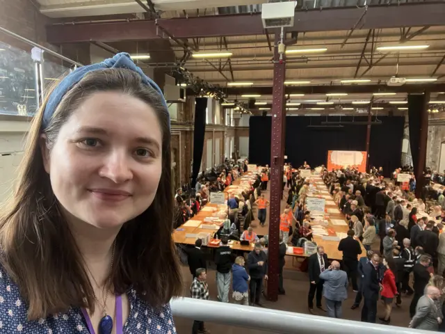 Reporter Sophie Parker selfie - she looks into the camera with a view of the counting from  a balcony - rows of trestle tables with people counting papers and others walking around watching the work.