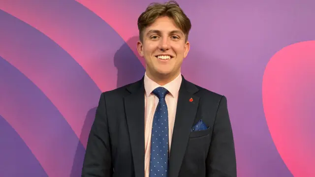 A 19-year-old smiles against a purple backdrop. He is wearing a navy suit and blue spotted tie and handkerchief and has brown hair.