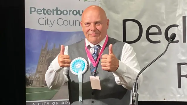 John Robert Bolton is standing on a stage in front of a Peterborough City Council sign which is on the left. He is wearing a white shirt