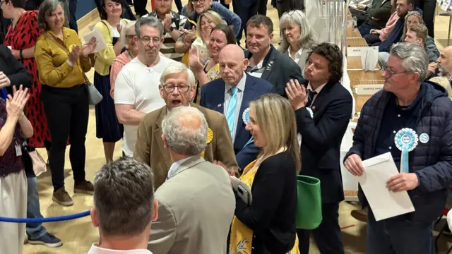 A group of candidates assembled at a small podium at an election count