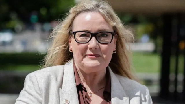 Karen Kilgour, who has long blonde hair and is wearing black glasses, a grey linen blazer and brown shirt, smiles at the camera.