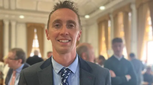 A man with short brown hair poses for the camera in a grey suit jacket over a blue shirt and dark blue tie weraing a political rosette.