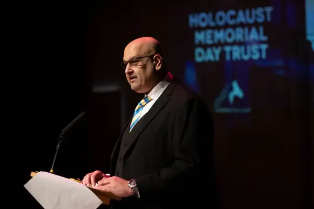 A man in a suit at a lectern. He is wearing a black jacket and waistcoast, blue and yellow striped tie and glasses as he talks into a microphone.
