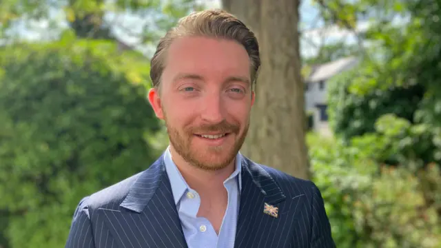A man in a navy striped suit and a flag pin on his lapel smiles at the camera