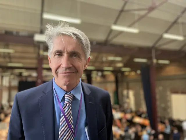 Paul Barltrop smiles at the camera - he wears a suit and tie and the count is blurred behind him in the background.