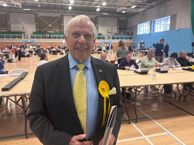 Garth Barnes, looking at the camera wearing a yellow tie with the count taking place behind him