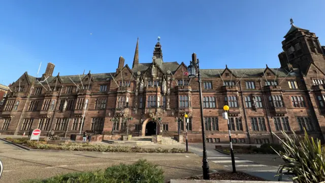 A view of Coventry's council house, a large four-storey building next to a zebra crossing. The sky behind is blue.