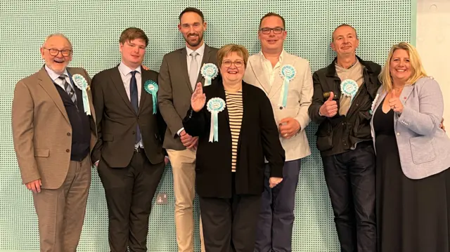 A group of seven Reform campaigners wearing light blue rosettes smile and give a thumbs up following success in the local elections