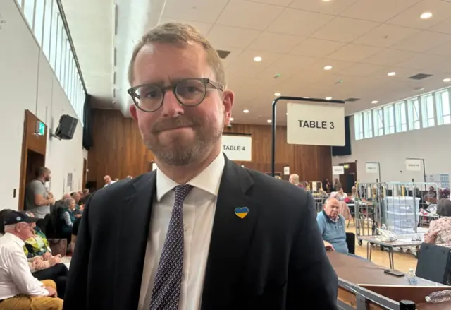 A man in a black suit with a yellow and blue heart pin and navy and white tie. He is standing in a hall with people at long tables, which are labelled with numbers, behind him. He has brown hair and dark-framed glasses, a neatly clipped beard and moustache.