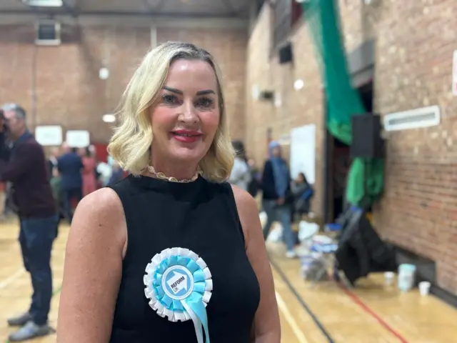 A woman with wavy blonde hair, and wearing a black sleeveless top with a blue and white Reform UK rosette pinned to it, smiles at the camera.