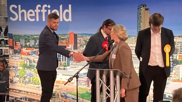 A man and woman shake hands in front of a blue background saying Sheffield