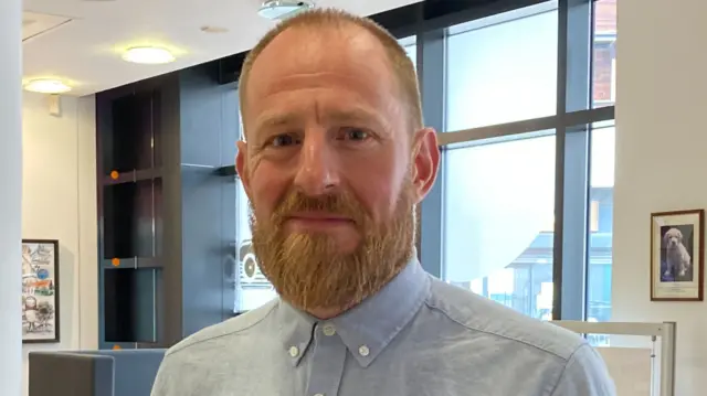 A man with a strawberry blond beard and hair in a blue shirt buttoned up to the collar. He is smiling in a reception area with white walls and large windows behind him.