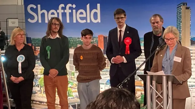 Six people stand on a stage. They are wearing political rosettes