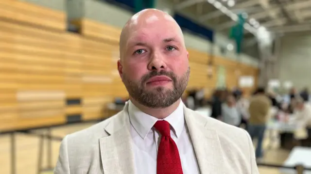 Bald man in white suit and red tie looking into thew camera. He is stood in a sports hall