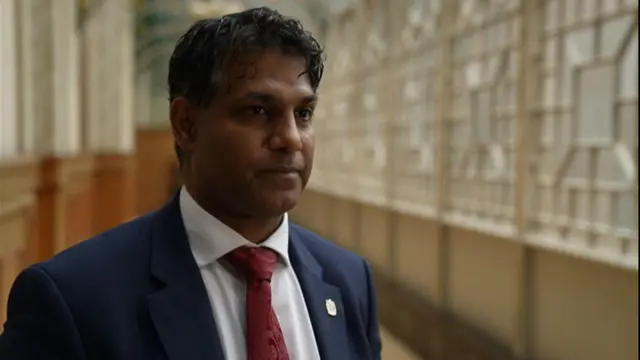 A man in a blue suit jacket, white shirt and red tie stands in a hallway with a neutral expression.