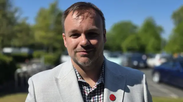 A man in a grey jacket and checked shirt smiles at the camera. He has short brown hair and a light beard