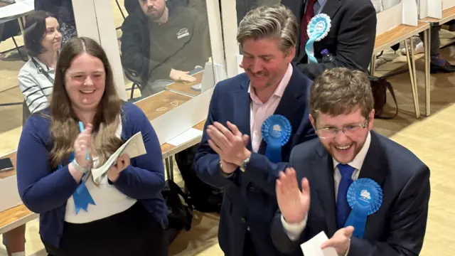 Three Conservative candidates, two men and a woman, applauding following the result in Welwyn East. A counter is sitting at a desk behind them.
