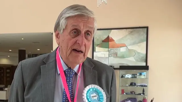 An elderly man in a suit with a blue Reform UK rosette on. He is giving an interview