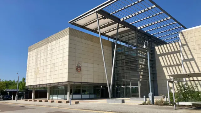 The outside of the South Cambridgeshire District Council building. It is a concrete cube-shaped building supported by concrete columns with a glass building in between covered by a metal canopy.