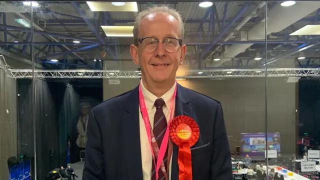 Sir Steve Houghton looks at the camera. He is wearing a navy suit, white shirt and dark coloured tie. He has a red Labour Party rosette pinned to his chest.