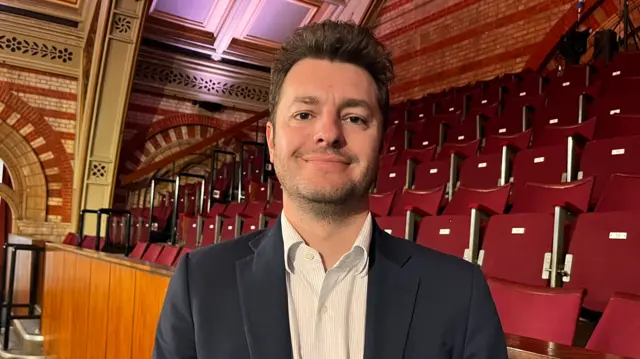 A man stands in front of seating area within a town hall. He smiles at the camera. He is wear a navy suit jacket with a white shirt underneath.