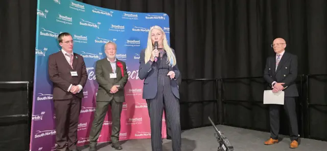 A woman with long white hair speaks into a microphone on a stage after an election result is announced, with two male candidates standing behind her and another man to her left.