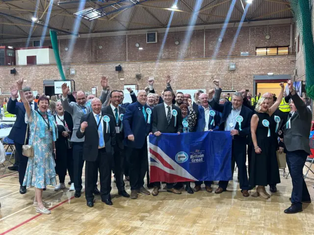 Large group of politicians celebrate and smile in front of a Reform UK flag