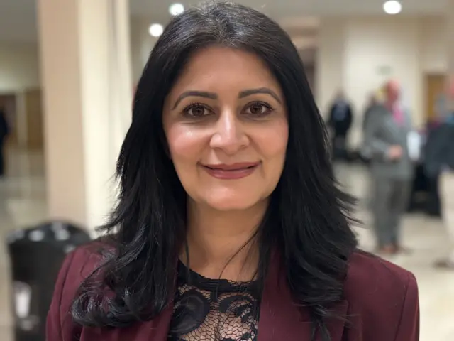 Shabina Qayyum is standing in the middle. She has long black hair and is wearing a black laced top with a burgundy blazer.