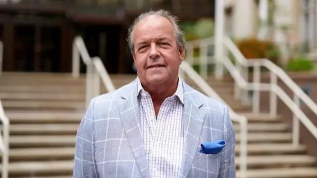 A man wearing a checked jacket and checked shirt looks at the camera. He is standing in front of some steps.