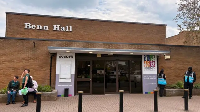 An external of Benn Hall's entrance, with people standing either side of a large glass door.