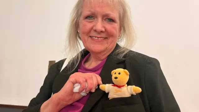A blonde woman in a black jacket and pink t-shirt holds the hand of a little yellow teddy tucked in her jacket pocket