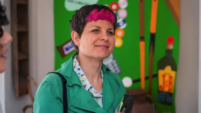 Zoe Garbett wearing green. she has pink fringe and a short, brown pixie cut.