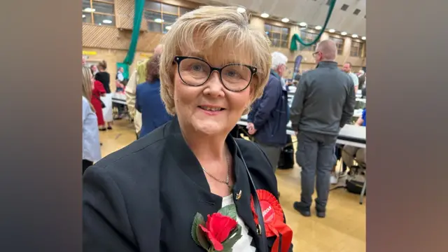 Tracey Dixon is smiling at the camera. She has short blonde hair and glasses. She is wearing a red rosette for Labour and a red rose on the lapel of her blue suit.