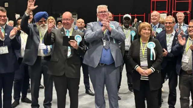 Two men and a woman in sky blue Reform rosettes celebrate, clapping and clasping their hands together as others around them applaud in a hall.